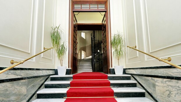 Uma escadaria imponente com tapete vermelho que conduz a uma porta, ladeada por duas plantas em vasos e corrimãos dourados.