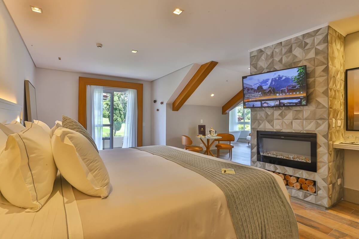Quarto de hotel moderno com uma cama grande, lareira, televisão e área de refeições com vista para o exterior.