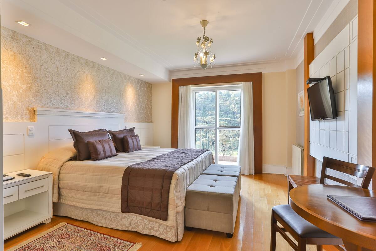 Quarto com uma cama grande, mesa de cabeceira, cadeira, mesa pequena, televisão e janela com vista para árvores.