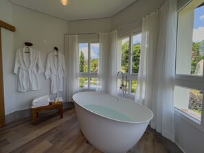 Ein modernes Badezimmer mit freistehender Badewanne, großen Fenstern mit Vorhängen und einer Holzbank mit Handtüchern.