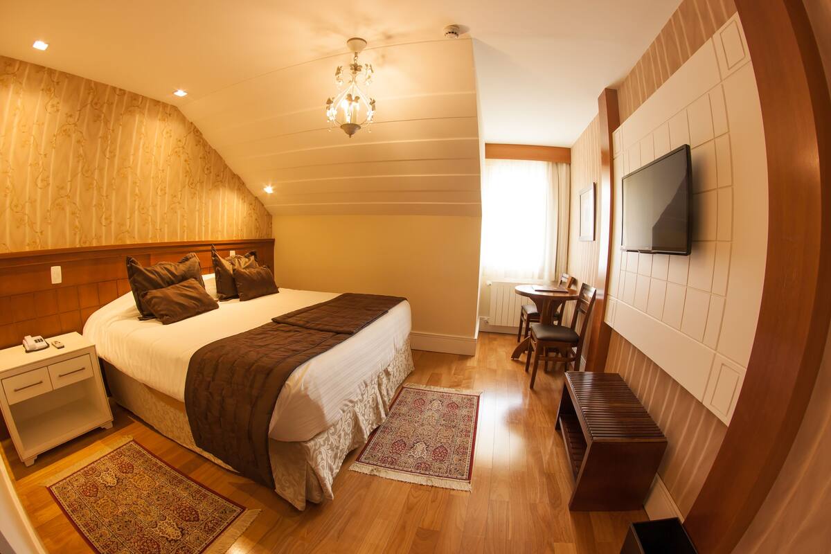 Quarto com uma cama grande, uma mesa de cabeceira, televisão na parede e um pequeno espaço de refeições com uma mesa e cadeiras.