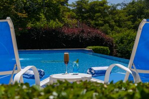 Ein Poolbereich mit zwei blauen Liegestühlen, einem runden Tisch mit einem Glas Champagner und einem blauen Getränk, umgeben von üppigem Grün.