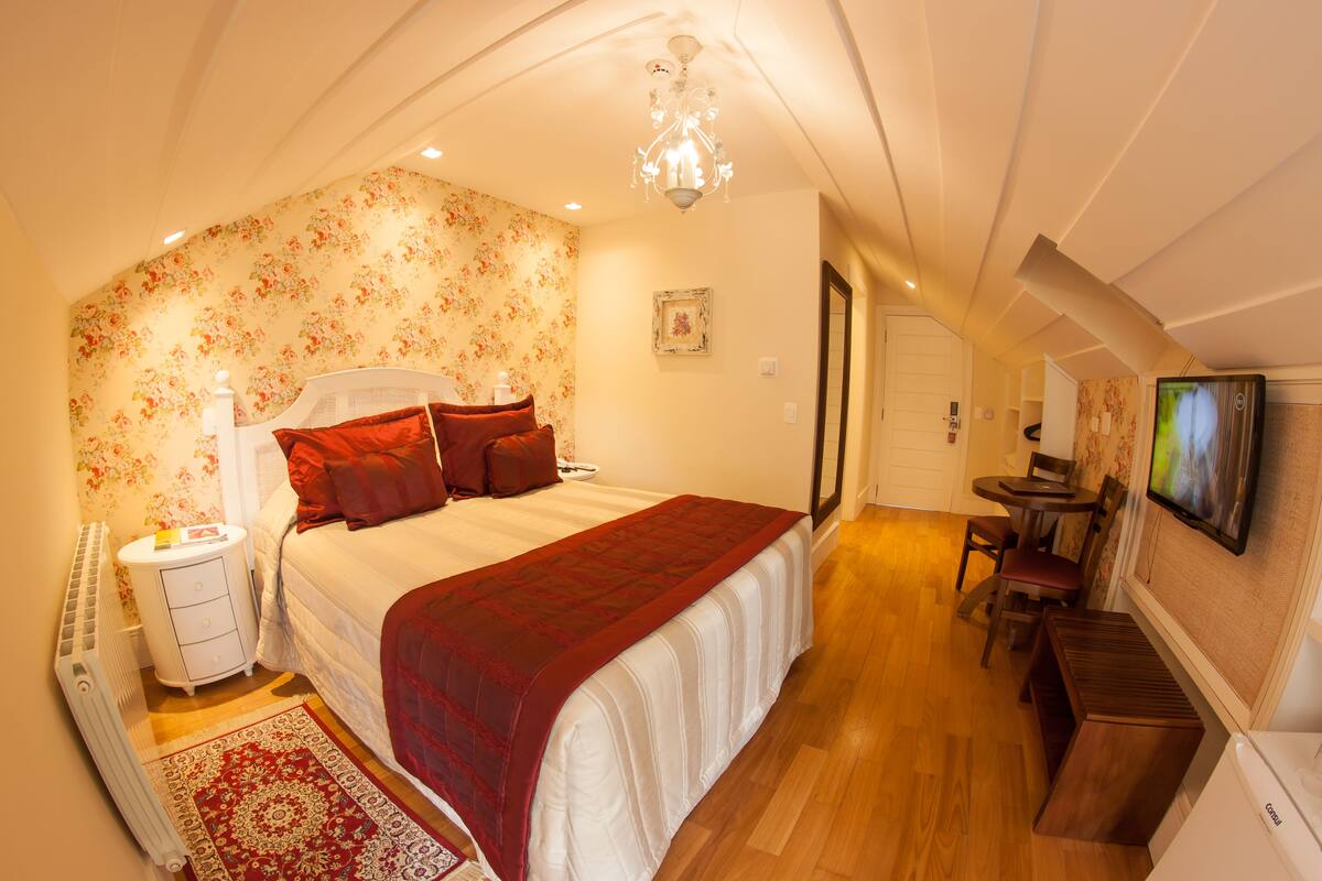 Quarto com uma cama grande, mesa de cabeceira, manta vermelha, piso de madeira, lustre, televisão e uma pequena área de refeições.