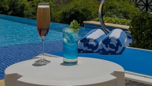 A poolside table with a glass of red wine and a blue drink, a blue and white towel, and a view of a hill with a building in the background.