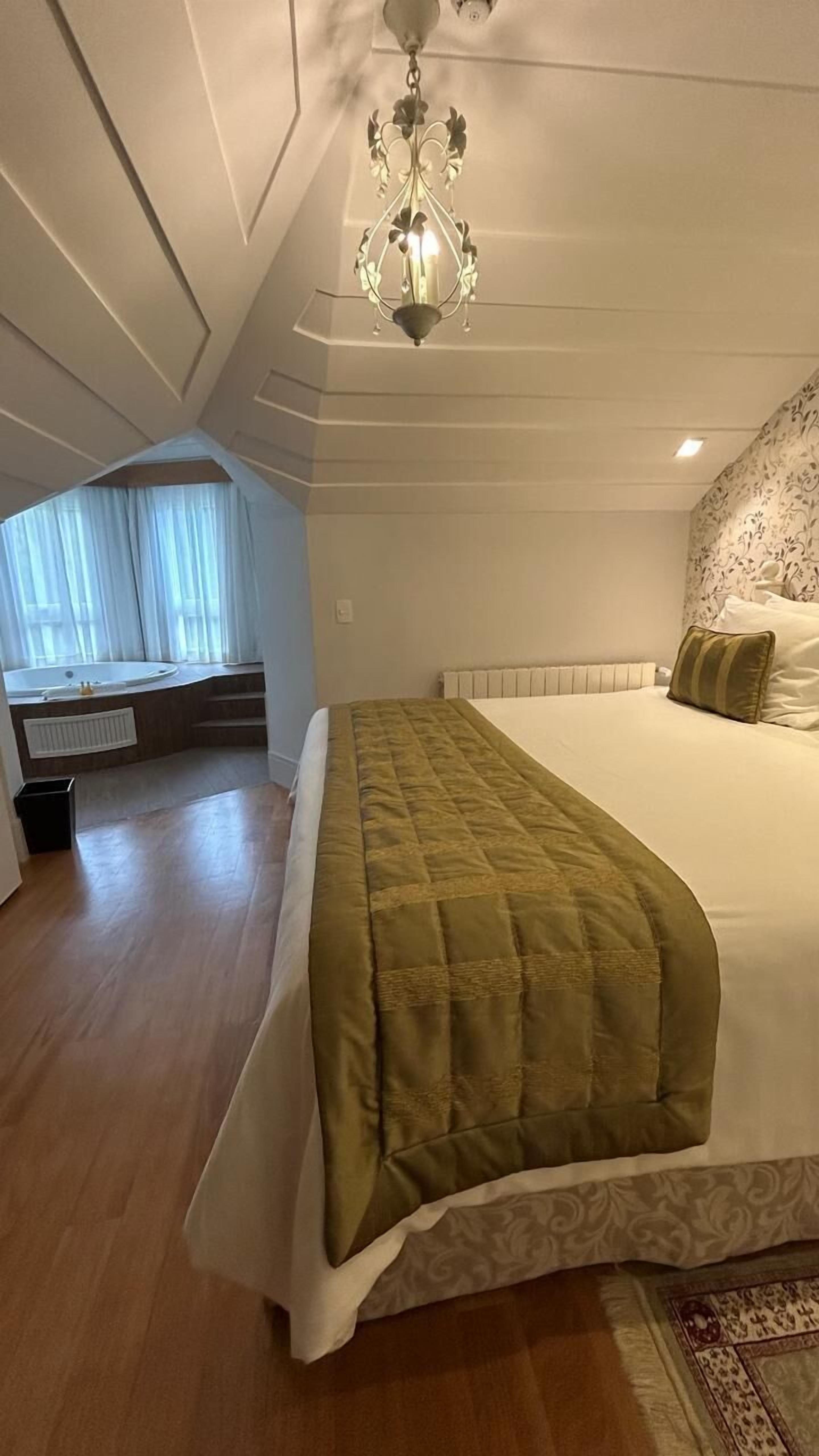 A bedroom with a large bed, a chandelier, a window with curtains, and a view of a bathroom.