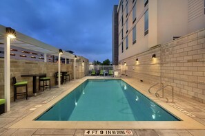 A rectangular indoor swimming pool with a depth of 4 feet 9 inches, surrounded by a tiled deck and a covered seating area with chairs and tables.