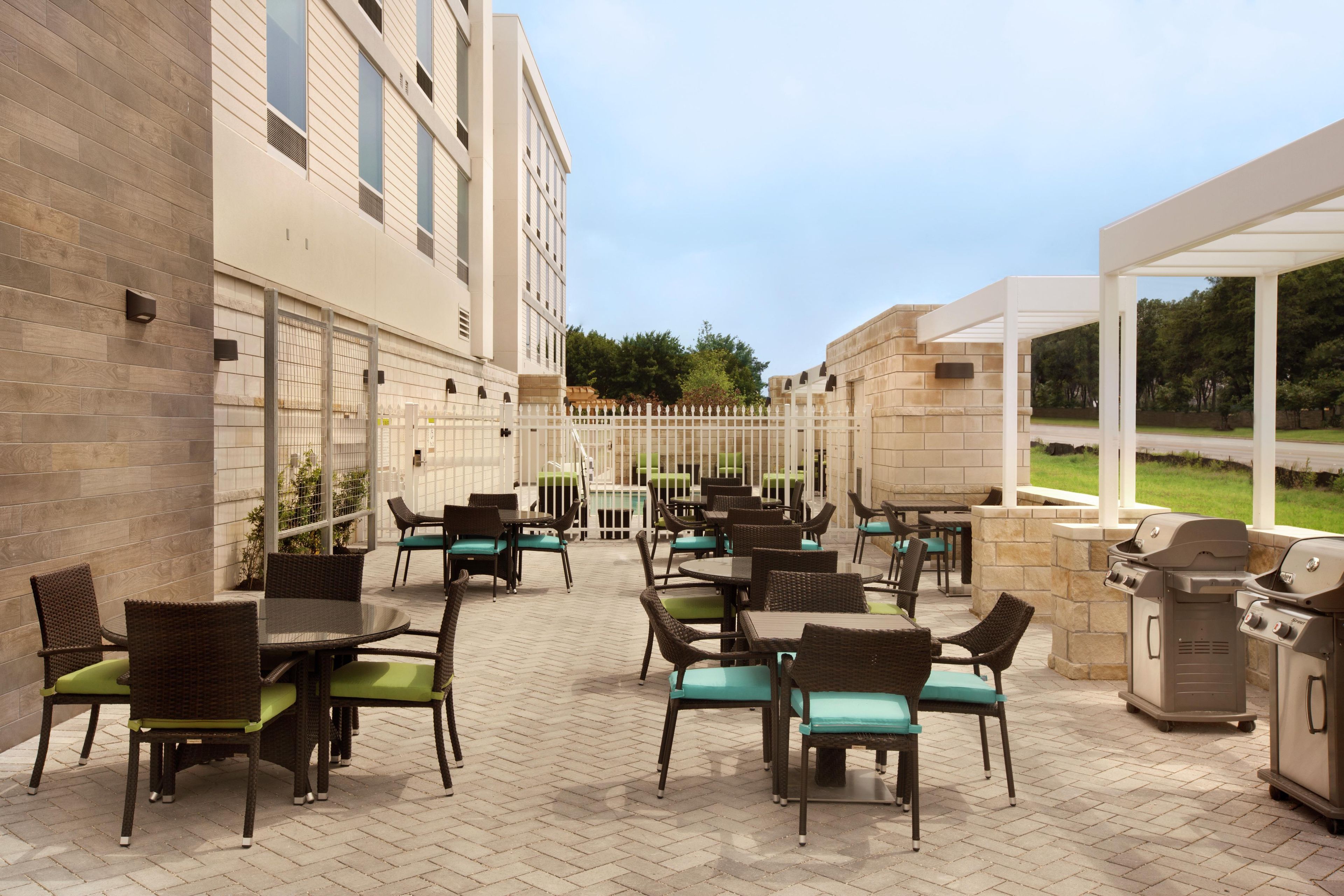An outdoor patio area with multiple tables and chairs, a barbecue grill, and a view of a building in the background.