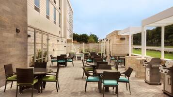 An outdoor patio area with multiple tables and chairs, a barbecue grill, and a view of a building in the background.
