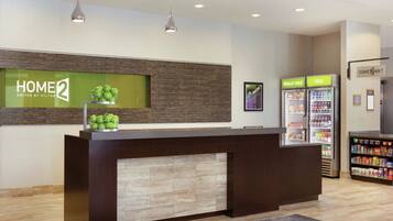 A reception area with a reception desk, a wall-mounted sign for 'HOME 2 SUITES BY HILTON', a 'REALLY COLD' refrigerator, and a beverage cooler.
