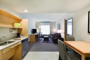 A compact hotel room with a kitchenette, a dining area, a seating area with patterned chairs, and a view of the outdoors.