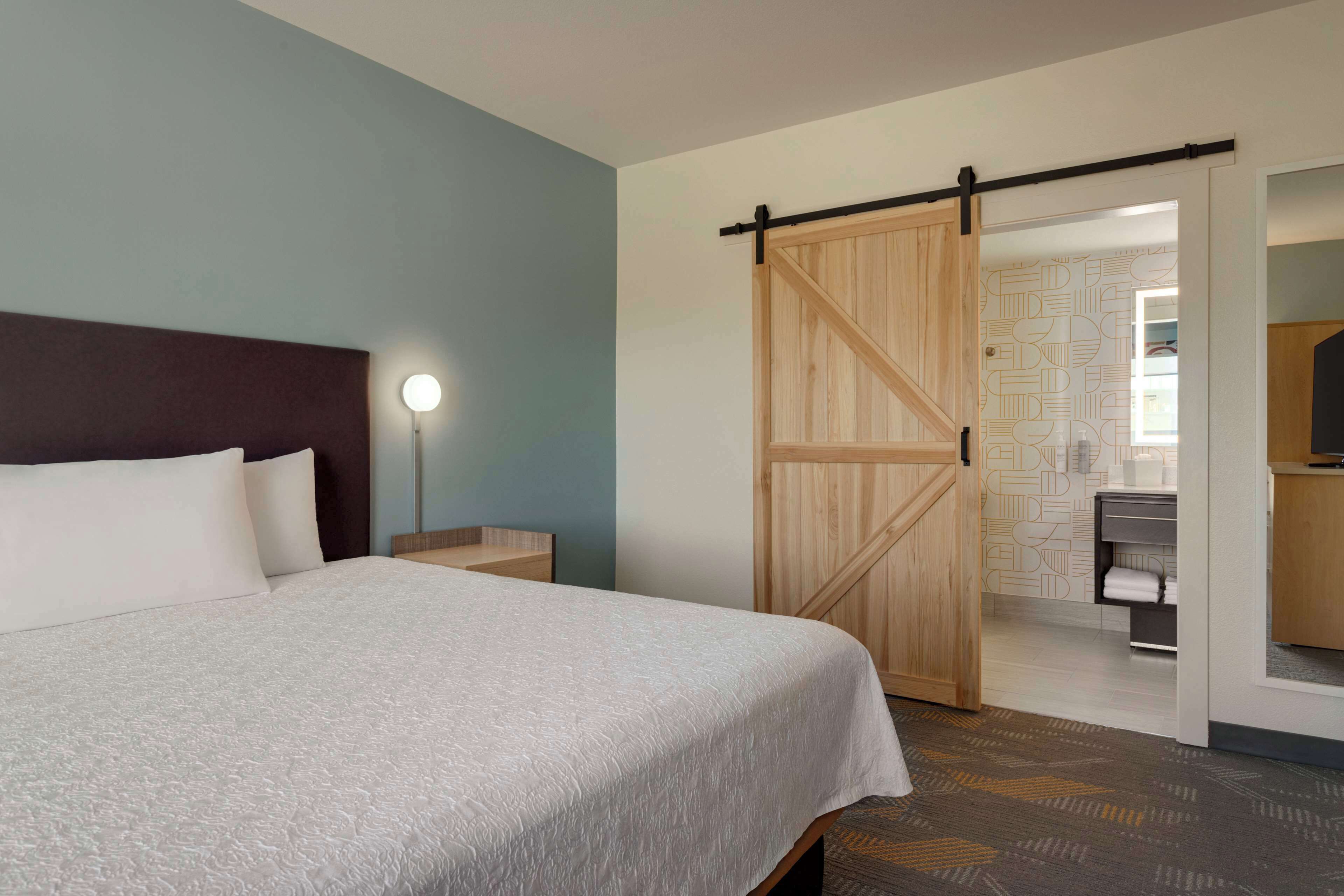 Una habitación de hotel con una cama, una mesita de noche, una lámpara de pared, una puerta corredera de madera y un baño visible a través de una puerta abierta.