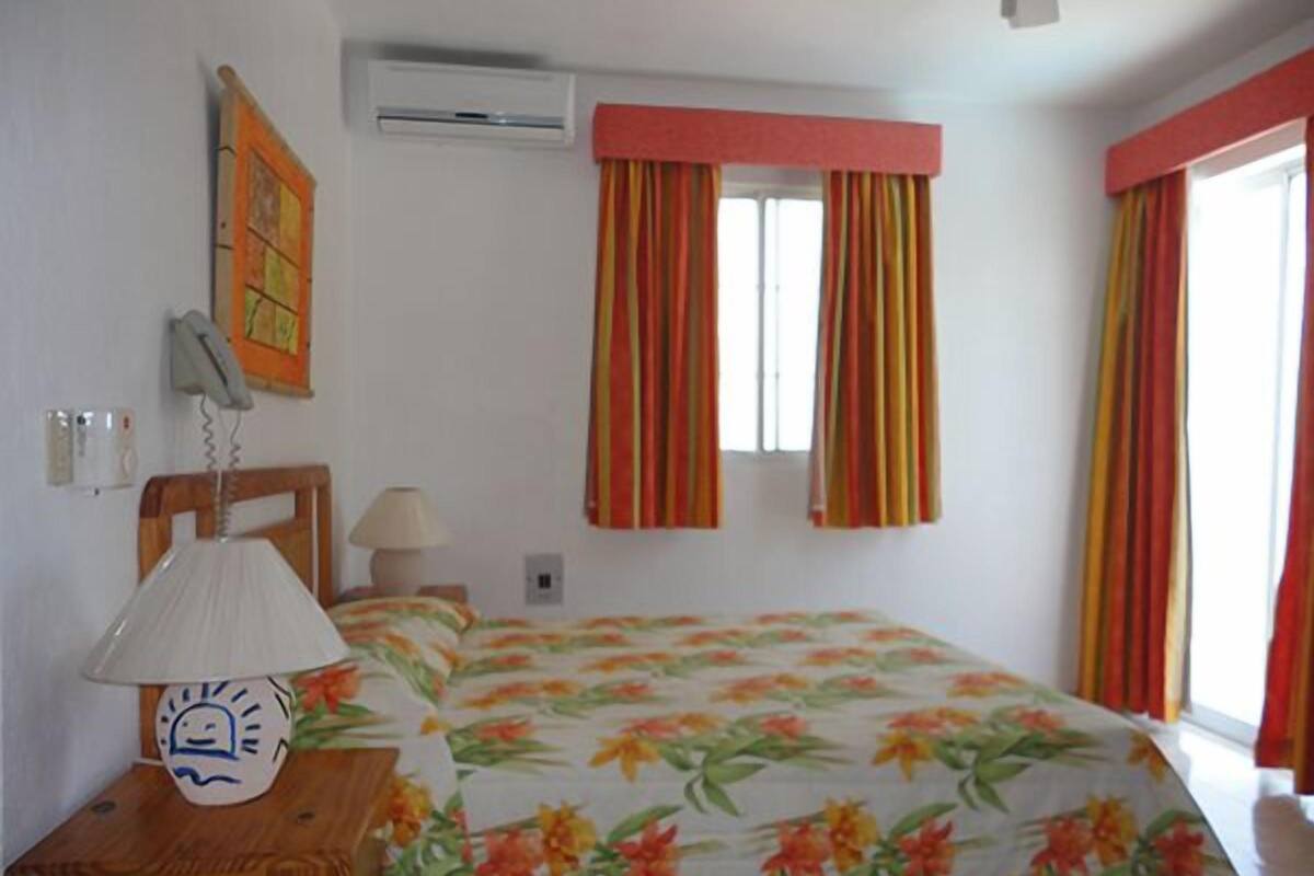 A bedroom with a bed, a bedside table with a lamp, a wall-mounted air conditioner, and striped curtains.