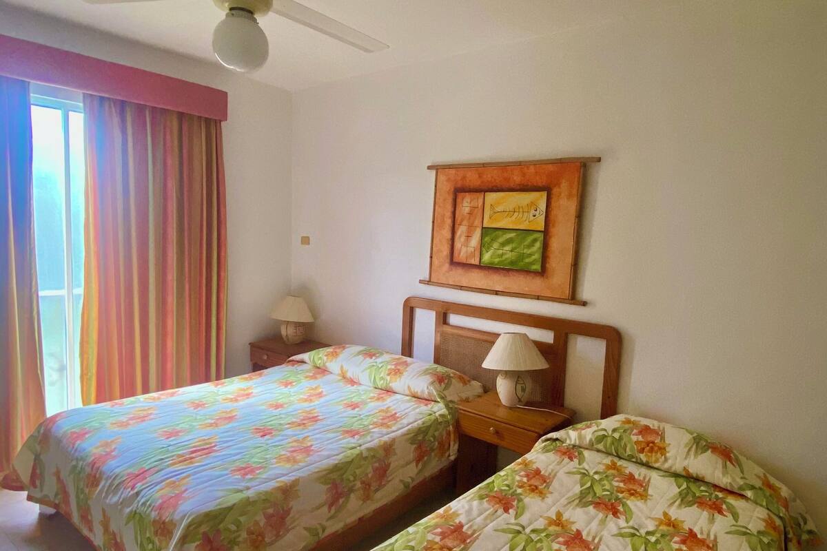 A hotel room with two single beds, a ceiling fan, a window with curtains, a nightstand with a lamp, and a wall decoration.
