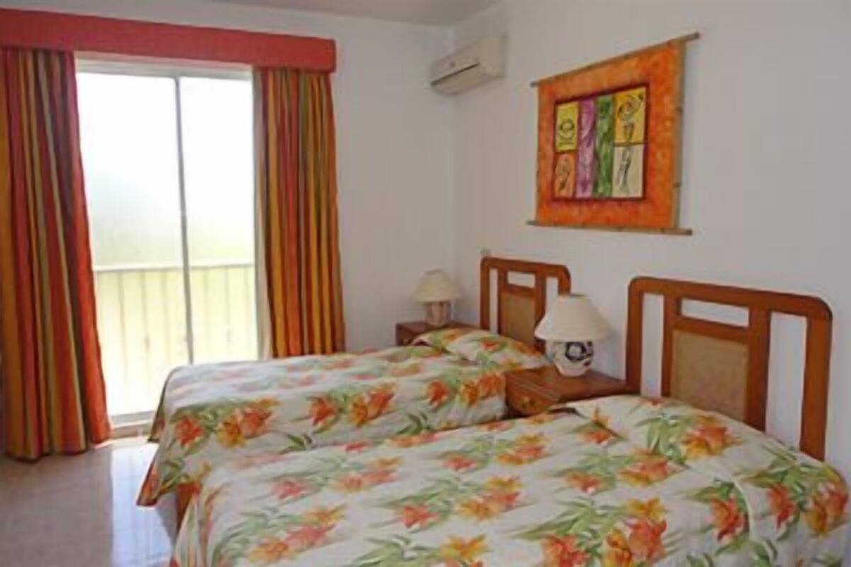 A hotel room with two beds, a window with curtains, a wall-mounted artwork, and a bedside table with a lamp.