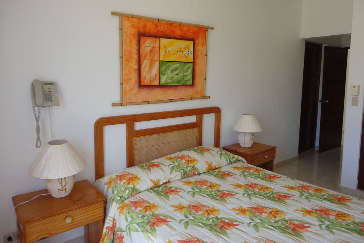 A bedroom with a bed, a bedside lamp, a telephone, and a painting on the wall.