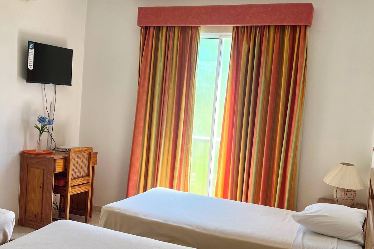 A hotel room with two beds, a desk with a computer, a TV mounted on the wall, and a window with colorful curtains.