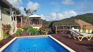 A rectangular swimming pool with clear blue water, surrounded by a wooden deck. There are lounge chairs and a covered seating area with a view of the surrounding greenery. - The Ocean Inn (English Harbour)