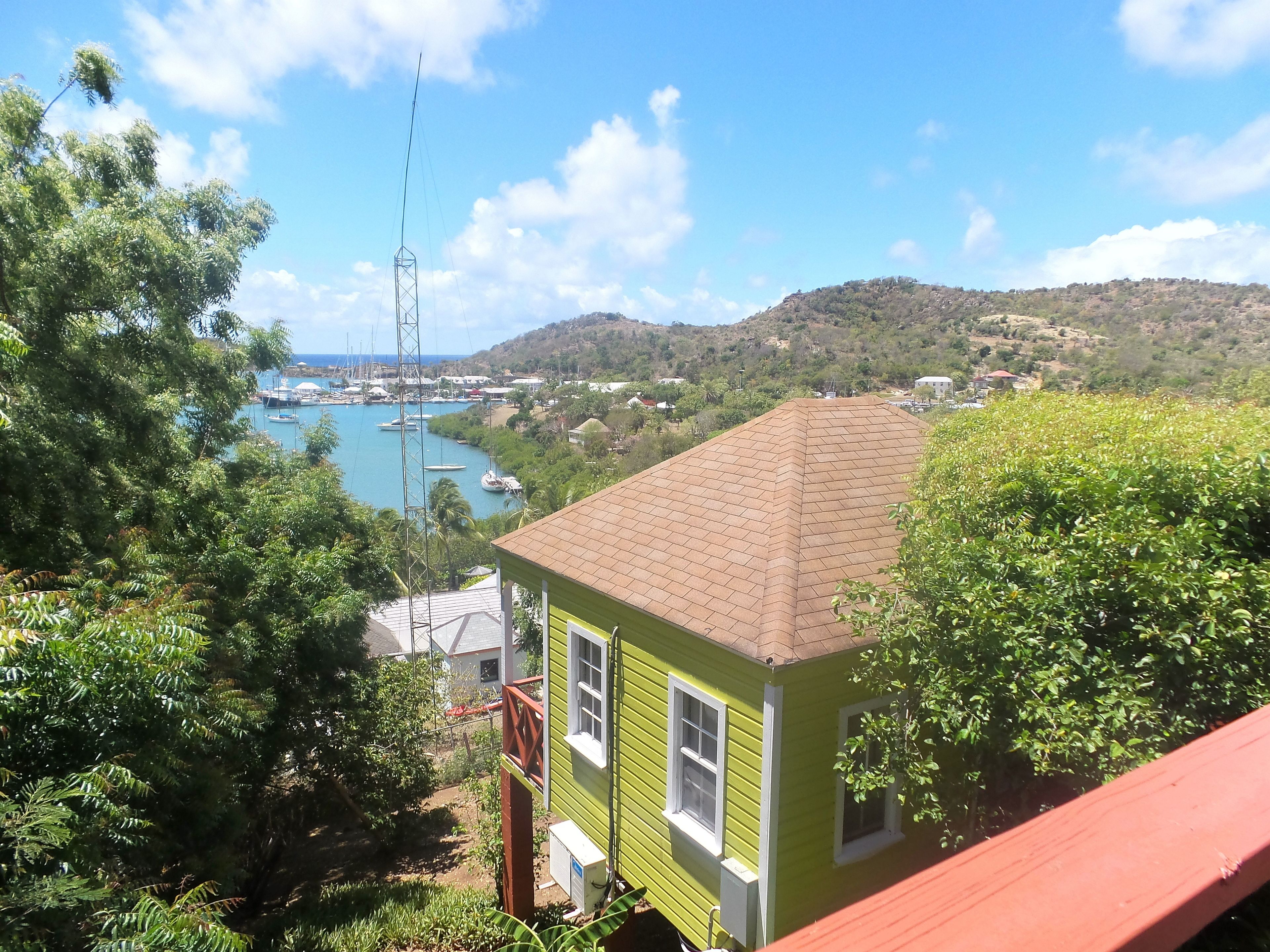 Um edifício verde com telhado ladrilhado, rodeado por vegetação exuberante e com vista para um porto com barcos.