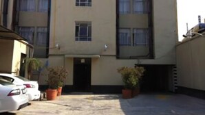 Un hotel de altura media con fachada plana, múltiples ventanas y un estacionamiento con varios coches y plantas en macetas.