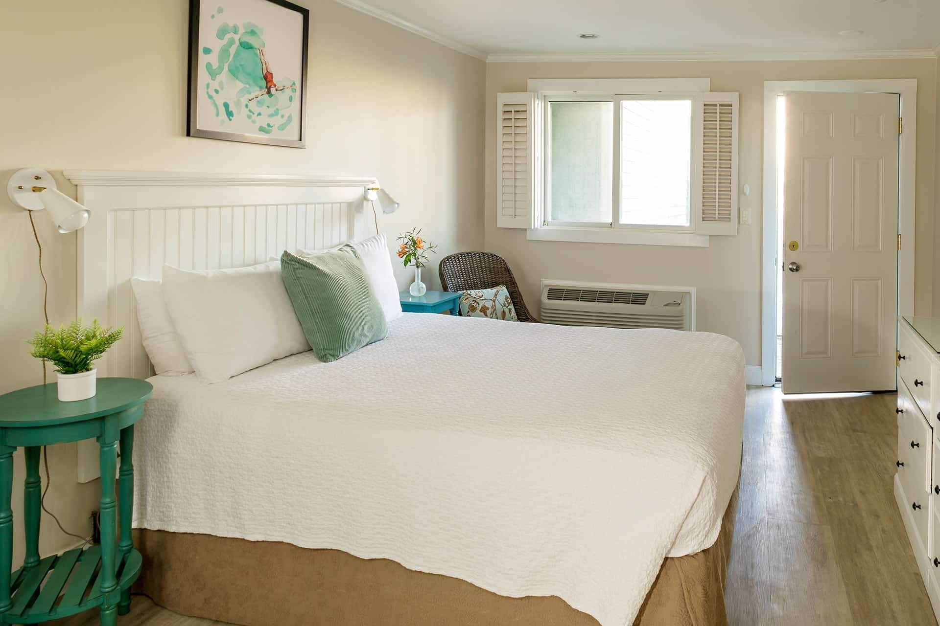 A bedroom with a bed, a nightstand, a lamp, a window with shutters, and a door.