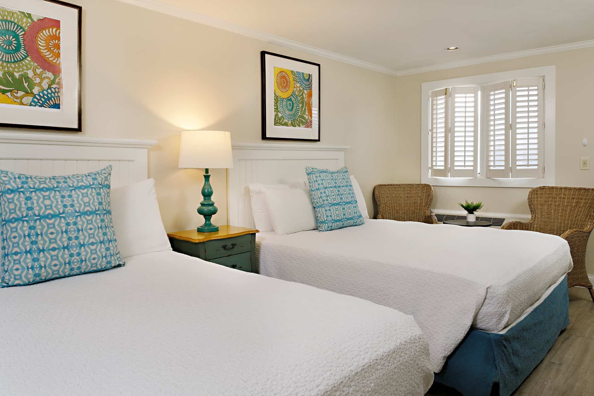 A hotel room with two beds, a nightstand with a lamp, a wicker chair, a small table with a plant, and a window with shutters.