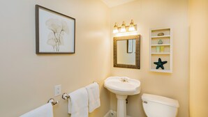 A bathroom with a pedestal sink, toilet, framed artwork, and a shelf with decorative items. - Madison Inn (Oak Bluffs)