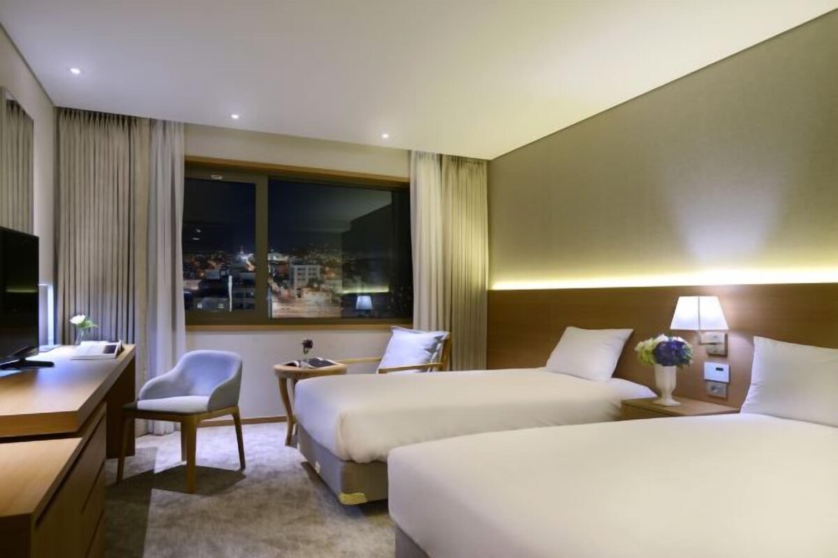 A hotel room with two beds, a desk, a chair, a TV, and a window with curtains.