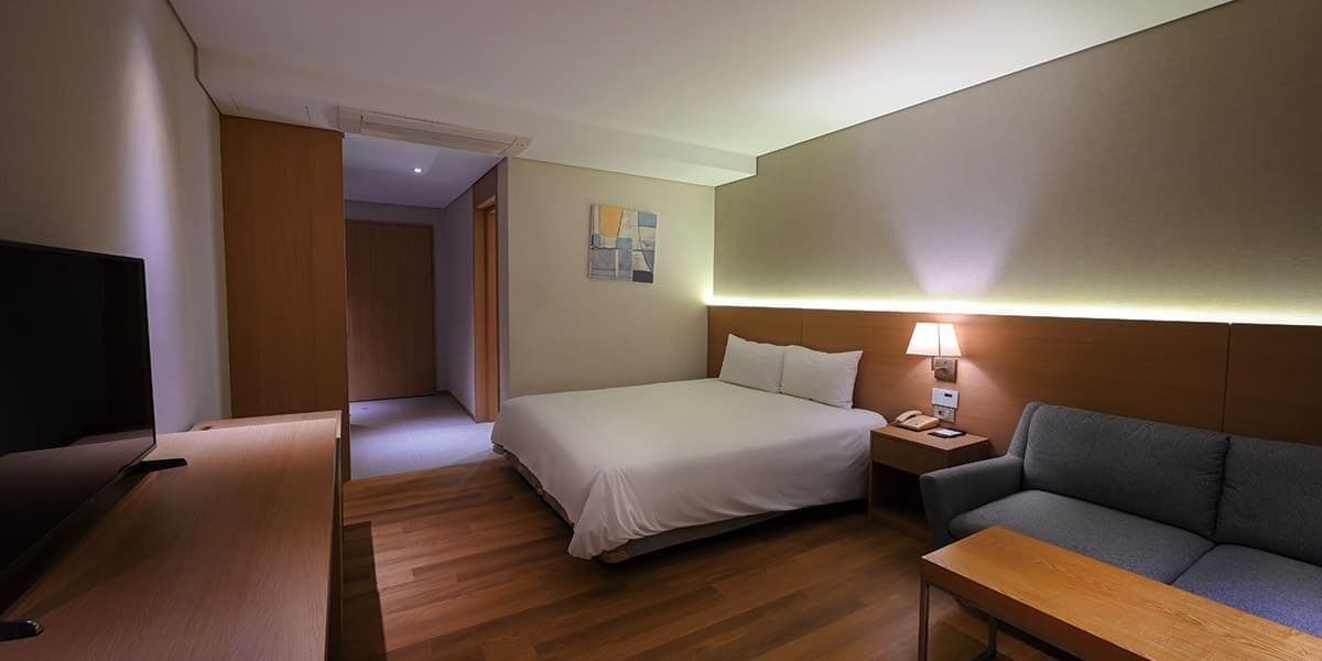 Quarto de hotel com cama, sofá, televisão, mesa de cabeceira e abajur.