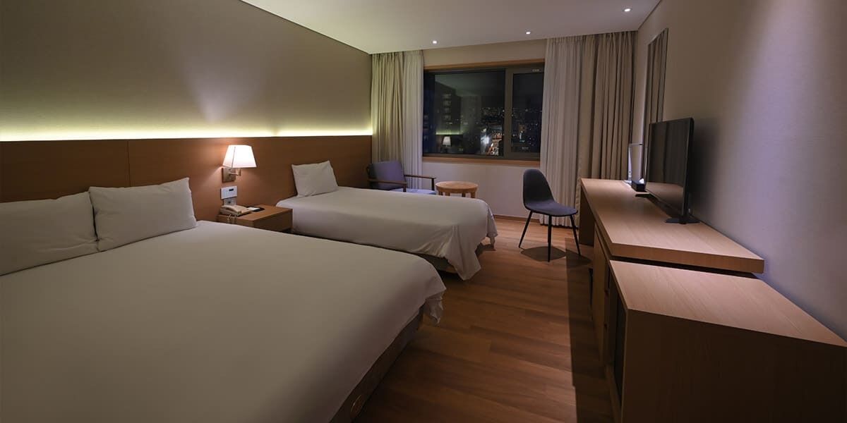 Habitación de hotel con dos camas, un escritorio, una silla, un televisor y una ventana con vista a la ciudad.