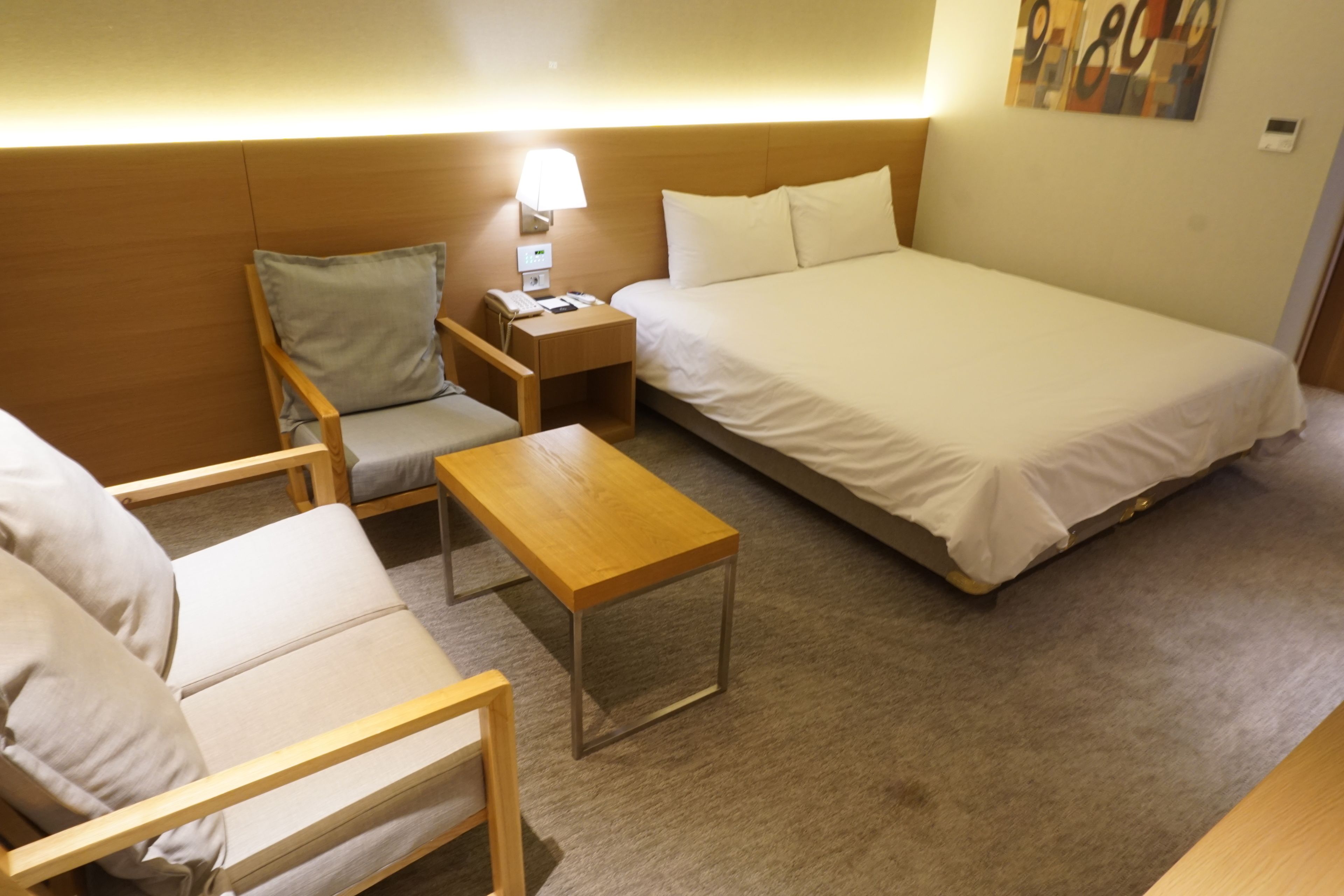 A hotel room with a bed, a chair, a small table, and a nightstand with a lamp.