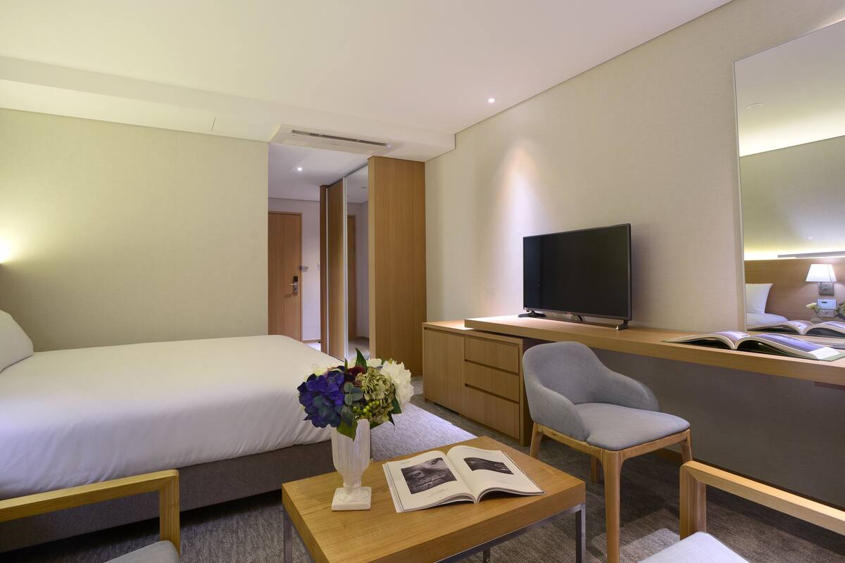 Habitación de hotel con cama, escritorio con televisor, silla, mesita con un libro y un florero.