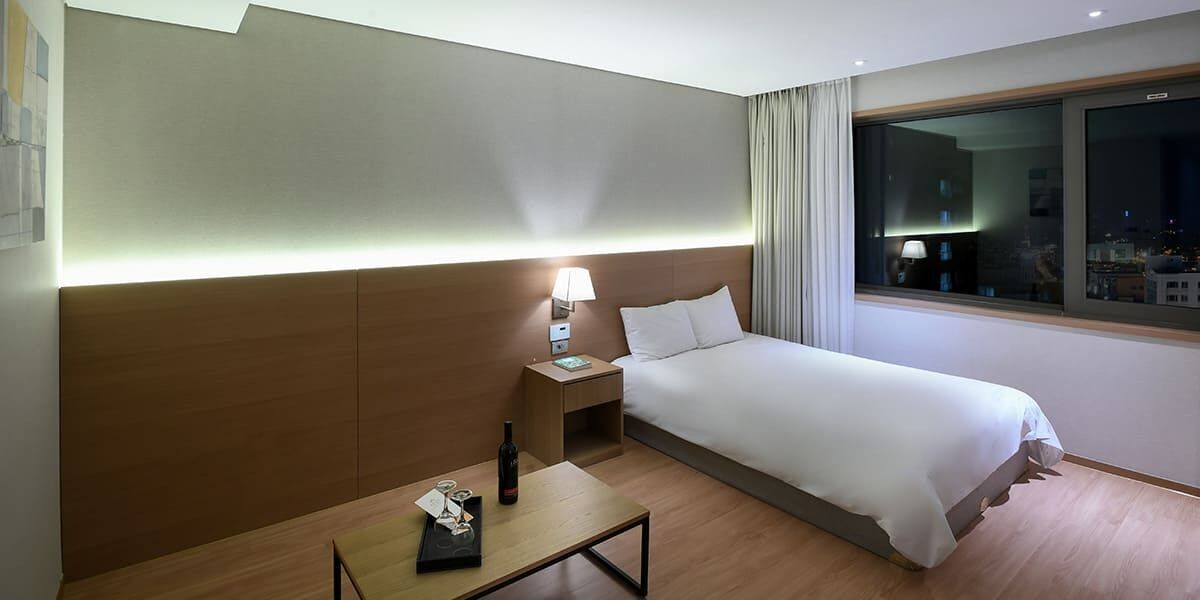 A hotel room with a bed, bedside table, lamp, and a window with a city view.