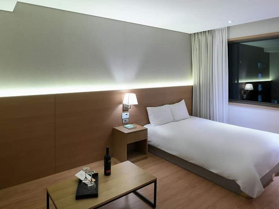 A hotel room with a bed, bedside table, lamp, and a window with a city view.