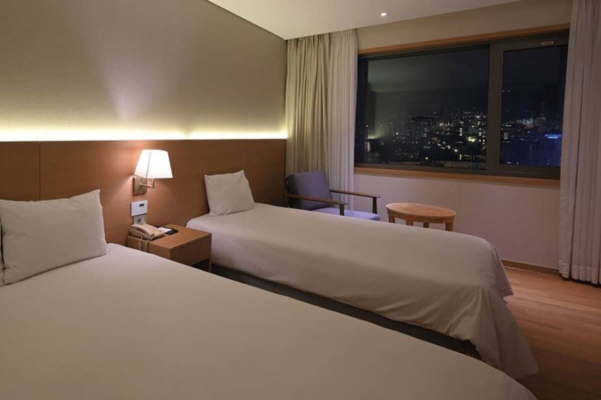 Habitación de hotel con dos camas, una mesita de noche con lámpara, una silla y vista a un paisaje urbano nocturno.