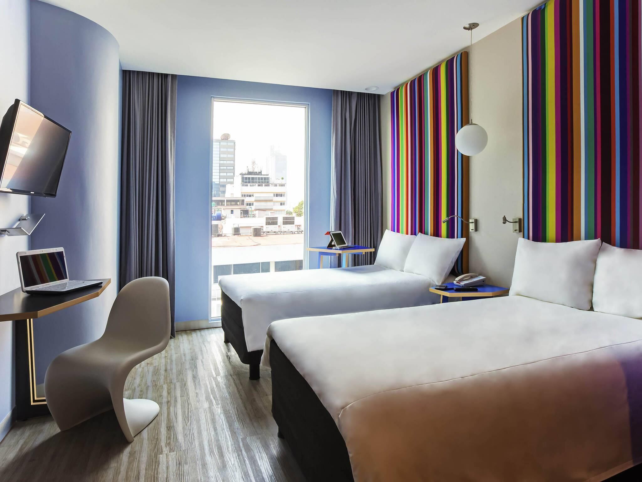 A hotel room with two beds, a desk with a laptop, a TV, and a view of the cityscape.