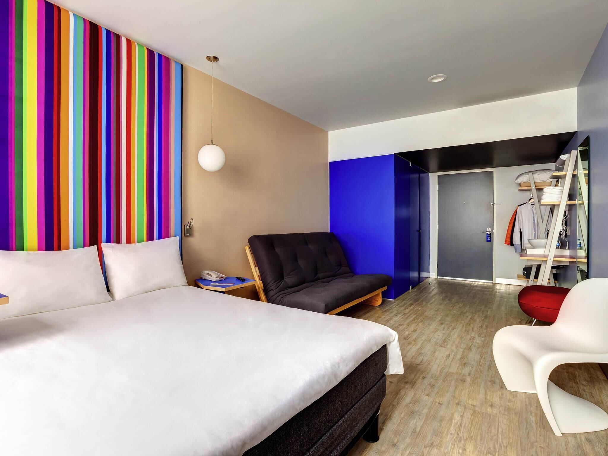 A modern hotel room with a bed, a black armchair, a white chair, a wardrobe, and a colorful striped wall.