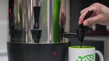 Una cafetera con diseño verde y negro, una persona sirviendo café en una taza blanca con un logotipo verde, y un letrero que dice 'CAFE Coffee'.