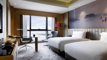 Habitación de hotel con dos camas, un escritorio, una silla, un ventanal con vista a la ciudad y un cuadro en la pared.