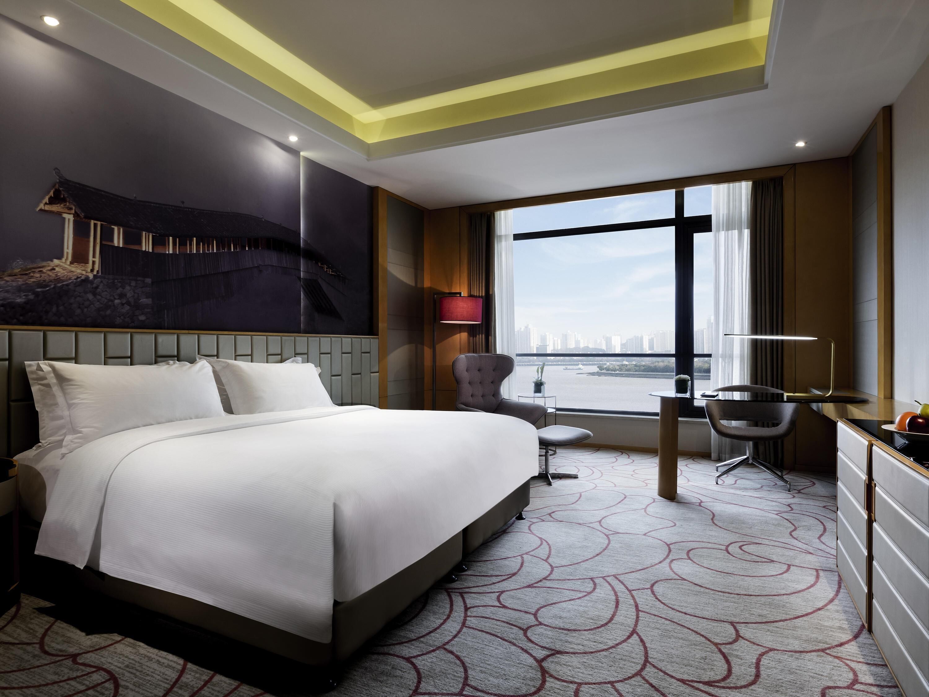 Habitación de hotel con una cama grande, un escritorio, una silla, vistas a un paisaje urbano y un diseño moderno de techo.