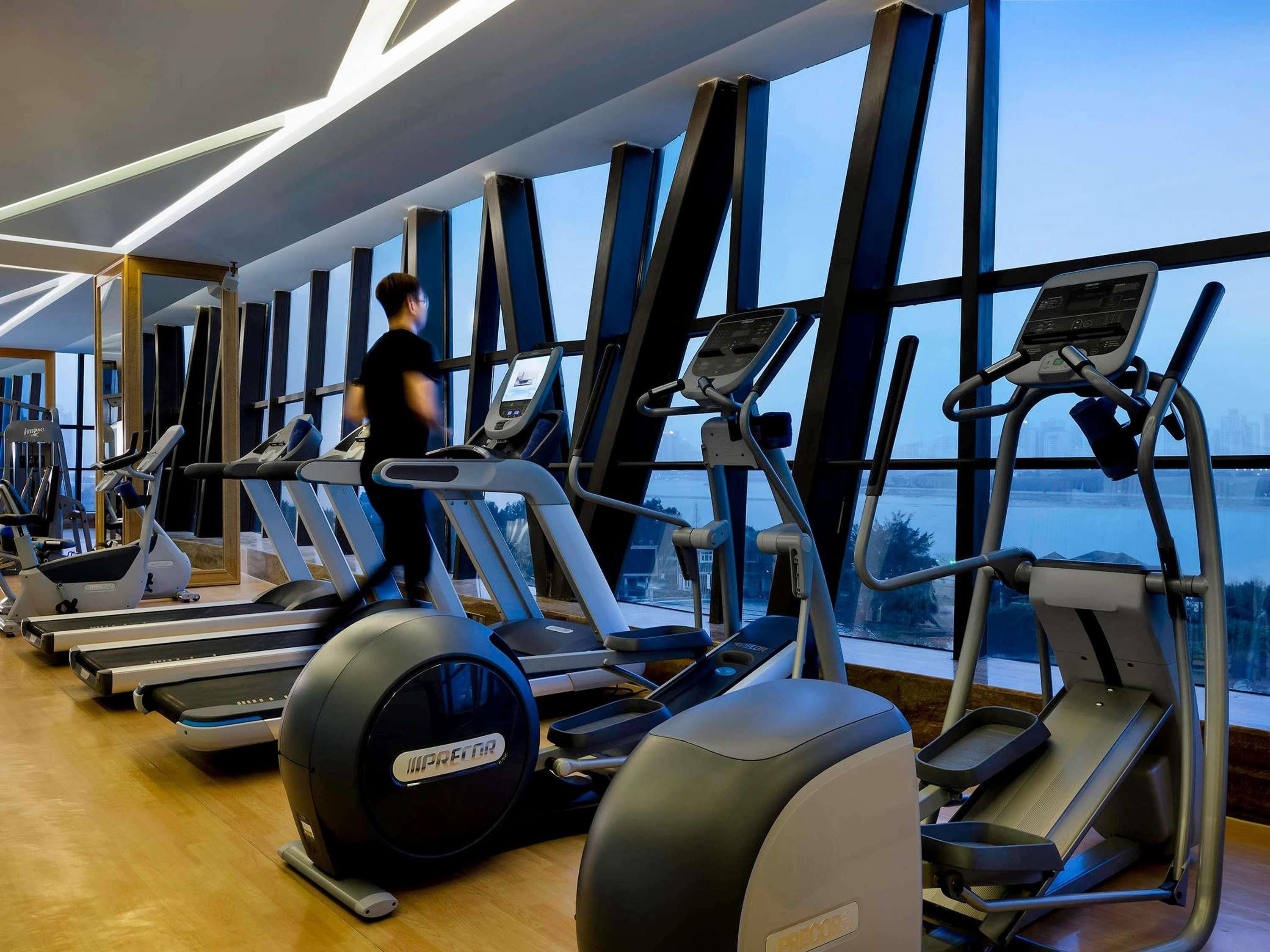 A gym with treadmills, ellipticals, and exercise bikes, large windows, and a person using a treadmill.