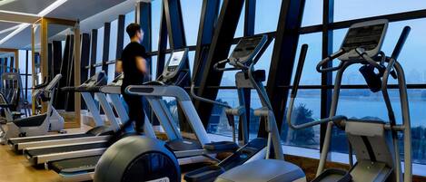 A gym with treadmills, ellipticals, and exercise bikes, large windows, and a person using a treadmill.