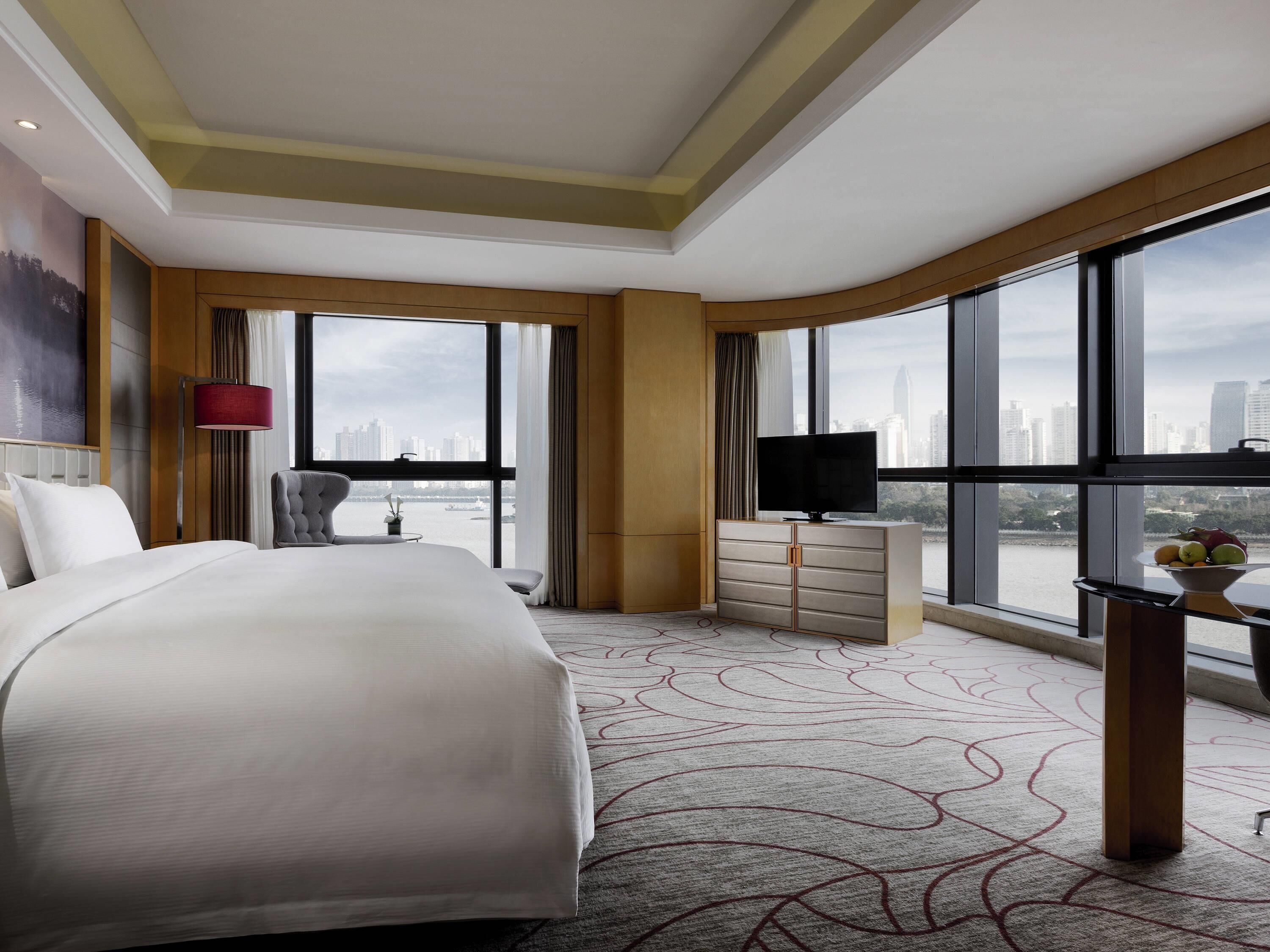 Habitación de hotel con una cama grande, televisión, escritorio con un frutero y vistas de la ciudad a través de amplias ventanas.