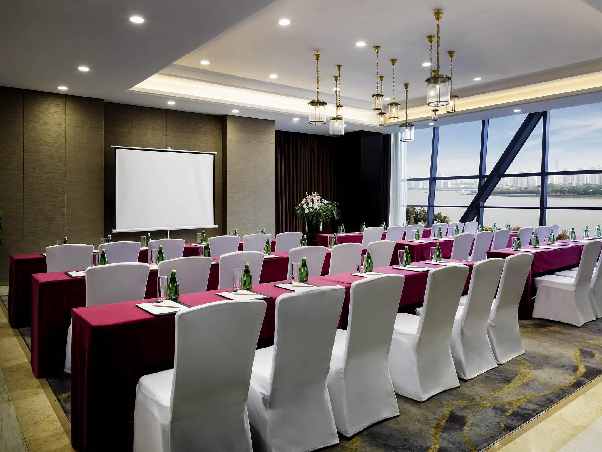 A conference room with tables set for a meeting, a projector screen, and a large window with a city view.