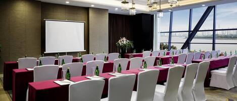 Un salón de conferencias con mesas dispuestas para una reunión, una pantalla de proyector y un amplio ventanal con vistas a la ciudad.