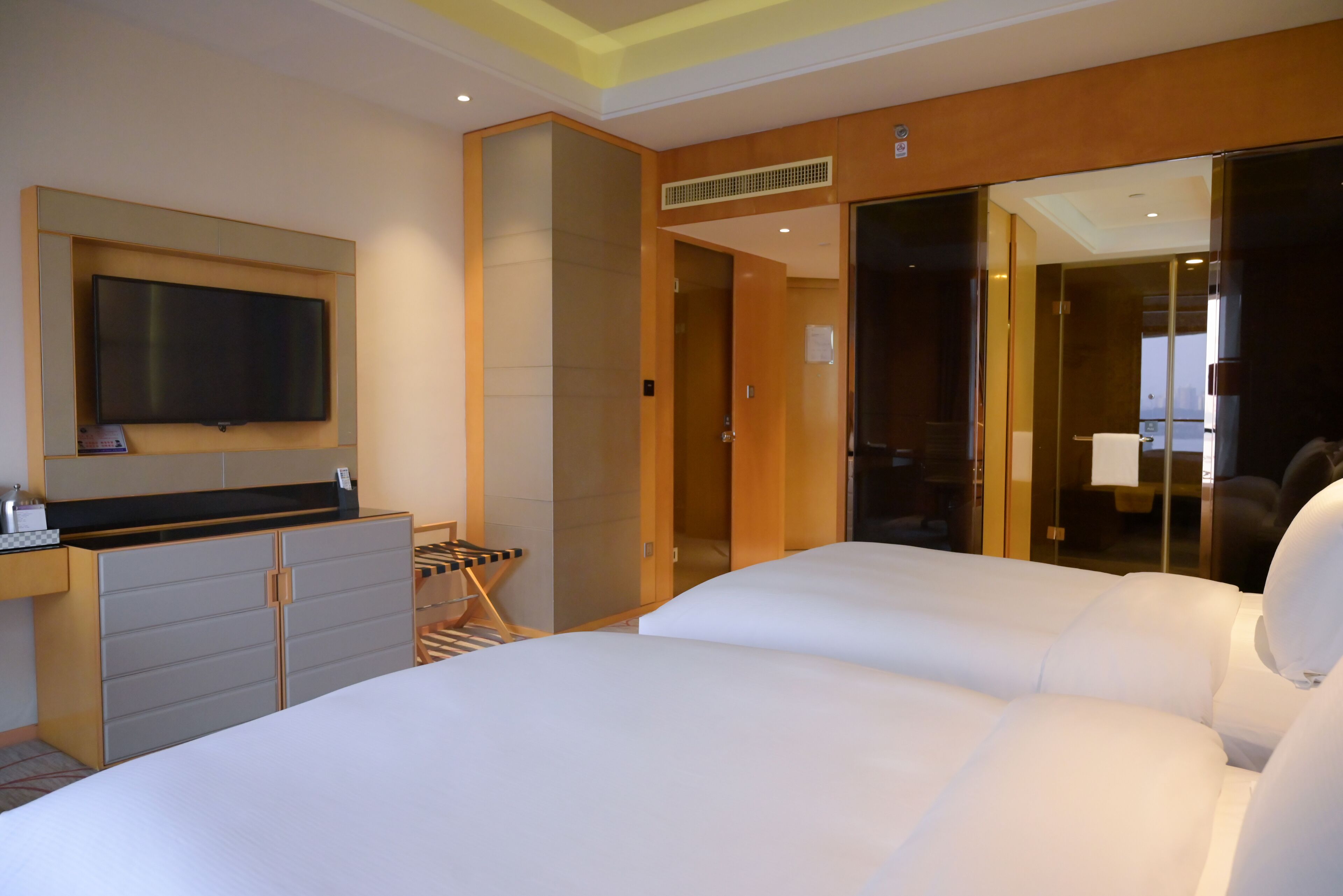 A hotel room with a large bed, a flat-screen TV mounted on the wall, a wooden wardrobe, and a sliding glass door leading to a bathroom.