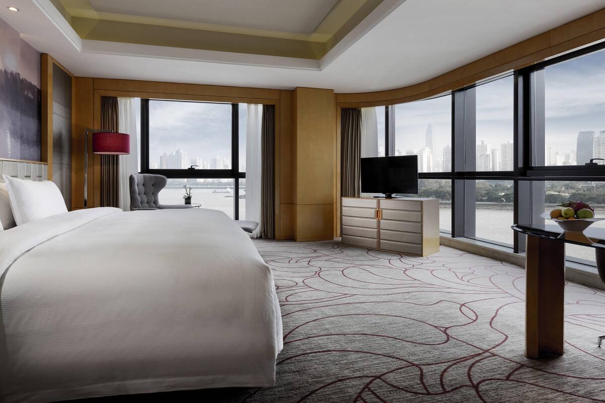 A hotel room with a large bed, a TV, a desk with a bowl of fruit, and a view of the city through large windows.
