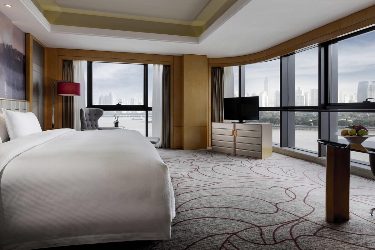 A hotel room with a large bed, a TV, a desk with a bowl of fruit, and a view of the city through large windows.