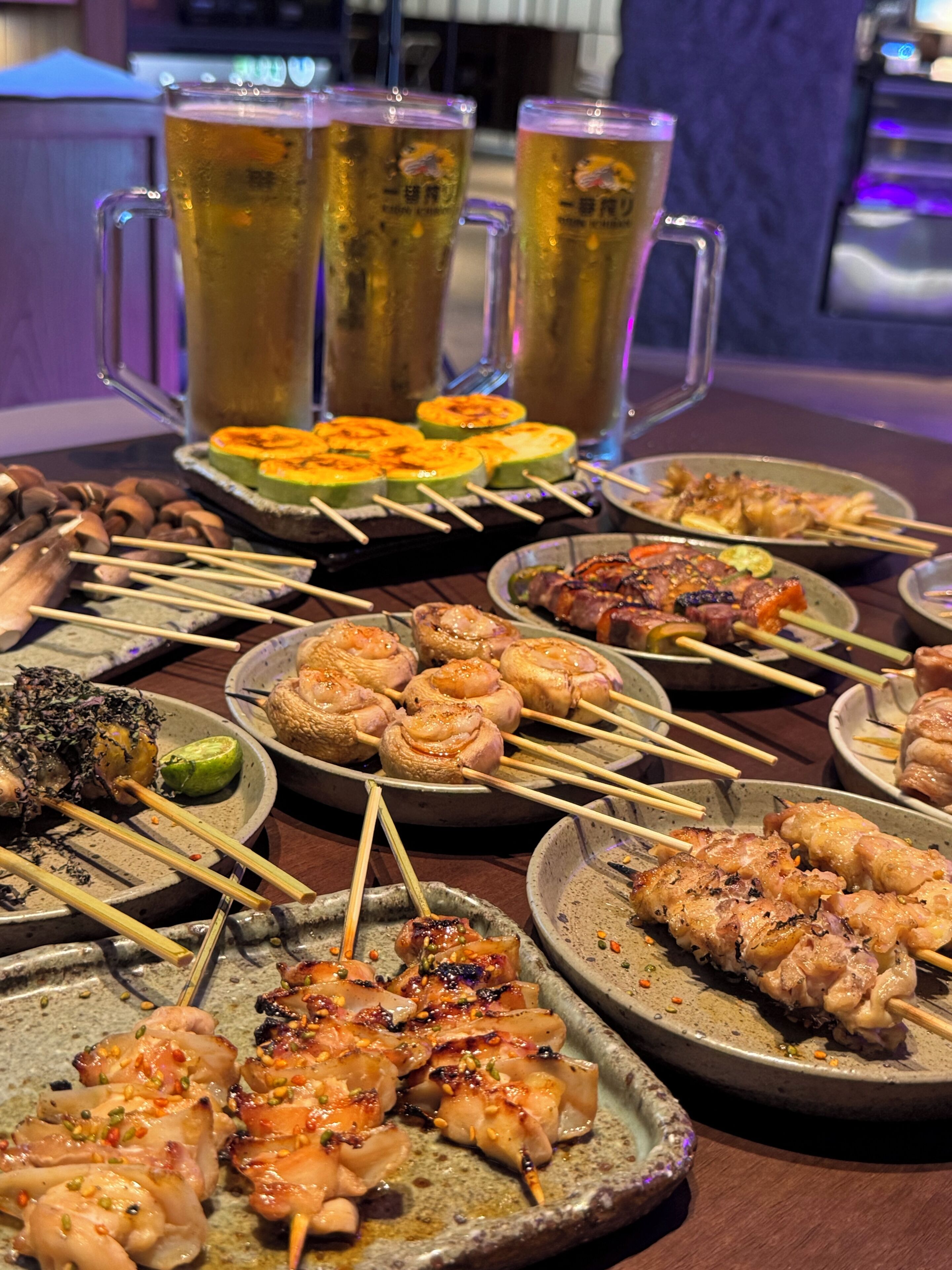 A variety of skewered meats and vegetables on ceramic plates, with beer mugs in the background.
