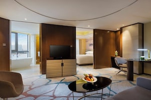 Habitación de hotel con una cama grande, televisión, escritorio, silla, una mesita con fruta y vistas a la ciudad a través de una ventana.