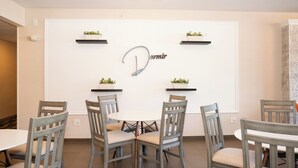 A dining area with a round table and six chairs, each with a wooden frame and cushioned seats. There are two black floating shelves above the table, each holding a small potted plant. The wall behind the table features a large, curved sign with the word "Dormir" and a decorative light fixture above it. - Dormir (Windham)